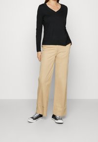 Svart V-ringad långärmad topp som kombineras med beige vida byxor och svarta sneakers, vilket visar en avslappnad passform och en mjuk tygstruktur.