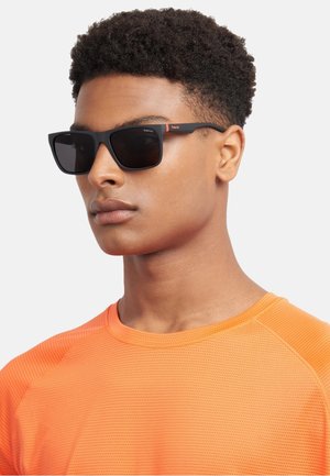 Occhiali da sole polarizzati neri con finitura opaca, montatura rettangolare e branding con accenti arancioni sulle tempie, indossati da un modello con una maglietta arancione.