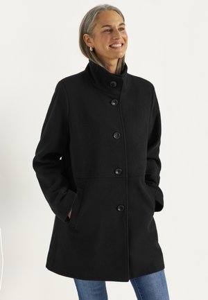 Kvinde med gråt hår iført en sort knap-et frakke med høj krave og blå jeans, smilende med hænderne i frakkelommerne.