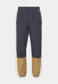 Sportovní kalhoty v námořnické modři a krémové barvě s pružným pasem a lemy, s hnědou spodní částí a bočními kapsami. Lehká, hladká tkanina.