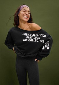 Felpa nera oversize con testo bianco che recita "URBAN ATHLETICS PLAY LOUD THE COLLECTIVE." Abbinata a leggings neri. Tessuto texture.