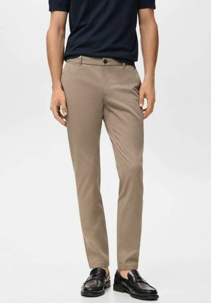 Pantalon beige coupe slim, confectionné dans un tissu lisse. Il présente une fermeture à un bouton et des poches latérales discrètes, associé à des mocassins noirs.