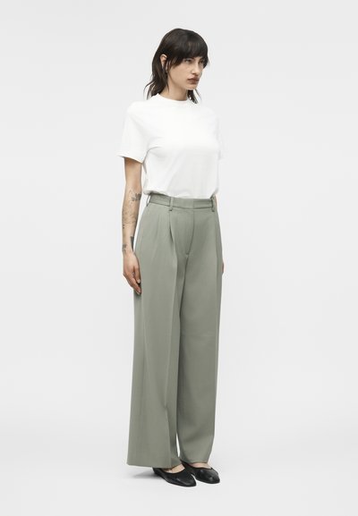 Γυναίκα με σκούρα μαλλιά στέκεται φορώντας λευκό κοντομάνικο τοπ, ψηλόμεσο παντελόνι φαρδύ σε ανοιχτό γκρι χρώμα και μαύρα επίπεδα παπούτσια μπροστά σε λευκό φόντο.