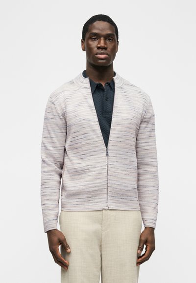 Homme portant une veste zippée gris clair sur un polo bleu foncé et un pantalon beige, debout devant un fond blanc uni.