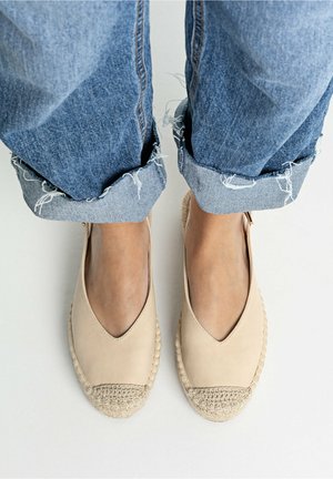 Pies con alpargatas beige con punteras tejidas y jeans azules deshilachados remangados sobre un suelo blanco.