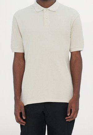 Mand iført en ensfarvet lyse beige kortærmet poloshirt med krave og to knapper, kombineret med sorte bukser, står foran en hvid baggrund.