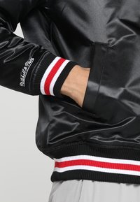 Sort satin bomberjakke med ribbet krave. Har røde og hvide stribede accenter på ærmerne. "Mitchell & Ness" logo broderet på ærmet.