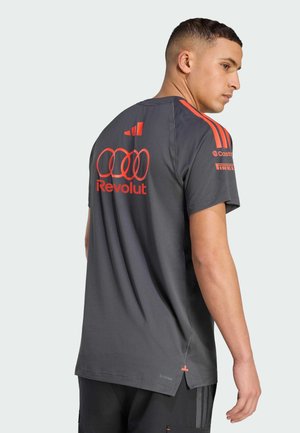 Uomo che indossa una maglietta sportiva grigio scuro con strisce Adidas arancione brillante e diversi loghi di sponsor, visto da dietro e di profilo.