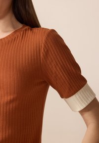 Ribētās adīšanas tops rūsas oranžā krāsā, ar īsām piedurknēm ar krēmkrāsas ribētām maliņām. Gluda tekstūra ar apaļu dekoltē un pieguļošu dizainu.