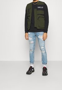 Person trägt eine olivgrüne Einsatzweste über einem schwarzen Langarmshirt, hellblaue, beschädigte Jeans, weiße Socken und schwarze Turnschuhe mit roten Details.