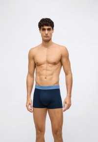 Young muscular man standing facing forward wearing navy blue boxer briefs with light blue waistband against plain background.