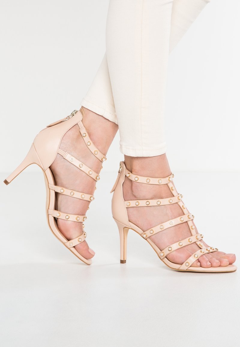 Pieds en sandales à talons hauts beige à brides avec des clous dorés, portées avec un pantalon blanc retroussé sur un fond blanc uni.