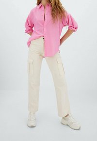 Camisa rosa de manga larga con botones, pantalones de cargo beige claros con bolsillos laterales y zapatillas blancas con suela texturizada. Fondo minimalist.