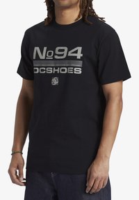 Svart bomullströja med fet vit grafisk text "No 94" och "DC SHOES", med korta ärmar och standard passform.