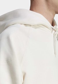 Felpa bianca in cotone con un ampio cappuccio strutturato, cordino e maniche raglan; tessuto testurizzato con dettagli di cucitura visibili.