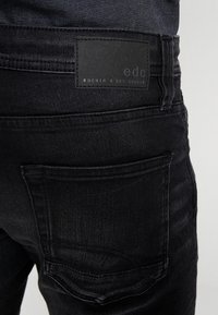 Jeans de mezclilla negra con un parche de cuero con la marca "edc" en la cinturilla trasera, diseño de cinco bolsillos y textura sutil.