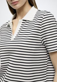 Πολο πουκάμισο με μαύρες και λευκές ρίγες, μαλακή υφή, κοντά μανίκια και γιακά με V γραμμή. Διαθέτει ομοιόμορφες οριζόντιες γραμμές.