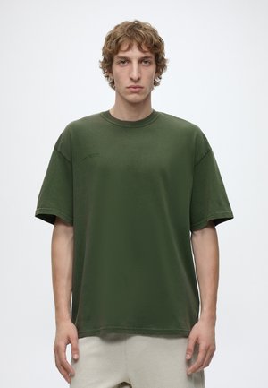 Junger Mann mit lockigem Haar, der ein übergroßes grünes T-Shirt und helle Hosen trägt und vor einem schlichten Hintergrund steht.