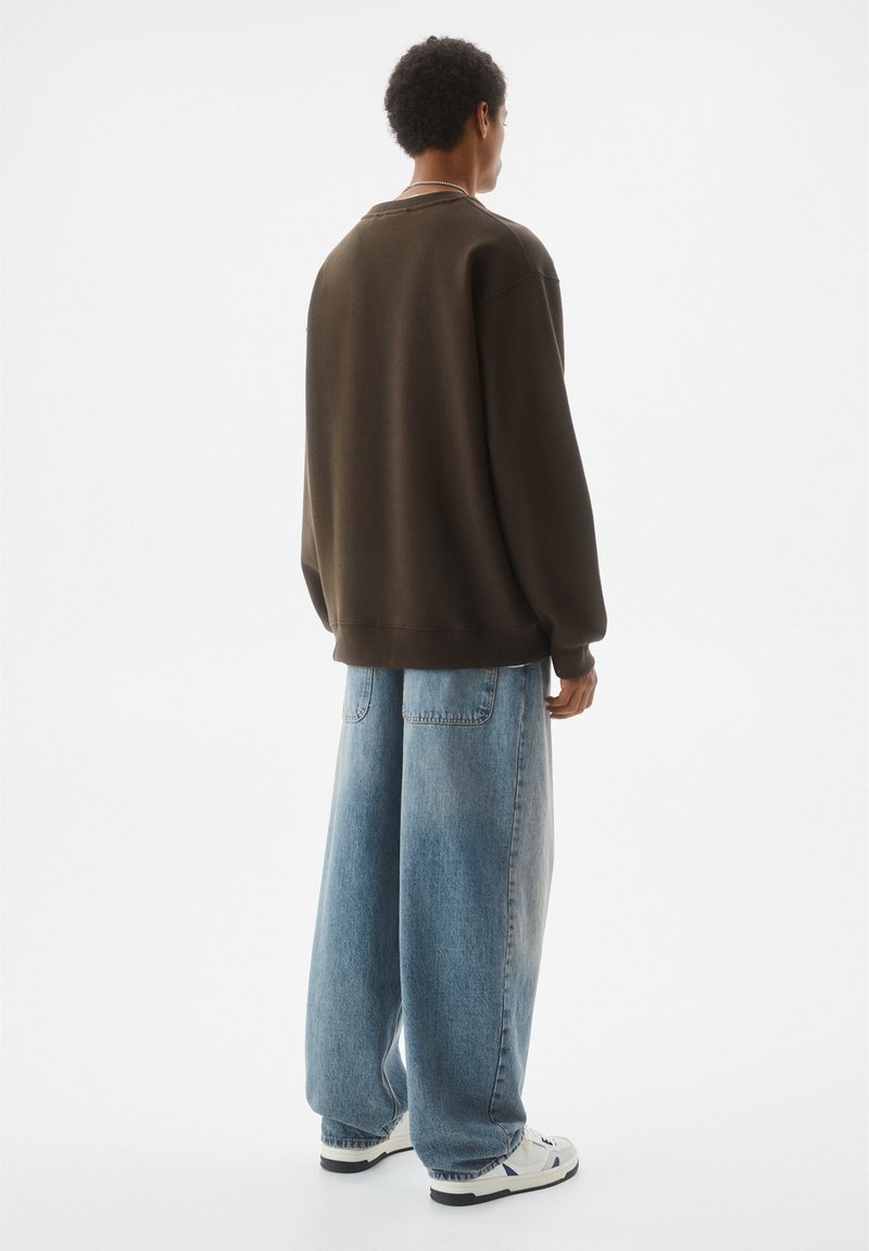 Homme aux cheveux courts et bouclés portant un sweat-shirt oversize foncé et un jean bleu délavé à jambes larges, debout de dos sur fond blanc.