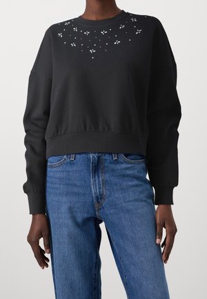 Zwarte cropped sweatshirt met lange mouwen en kleine bloemvormige uitsnijdingen bij de halslijn, gecombineerd met een hooggetailleerde blauwe spijkerbroek.
