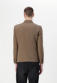 Blazer marrón a medida con un ajuste estructurado, abertura sencilla y puños abotonados. Textura suave y silueta moderna. Jeans negros visibles abajo.