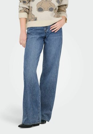 Kvinne iført blå vide bukser, beige genser med bamsemotiv, svarte spisse sko, stående med én hånd på hoften.