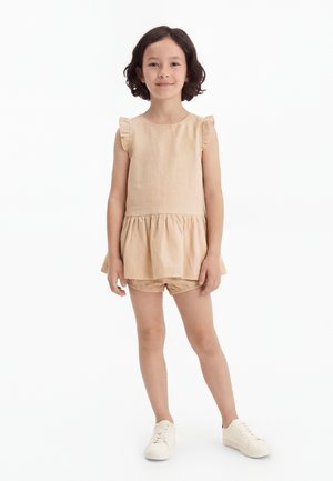 Jeune fille aux cheveux courts et foncés portant un haut beige sans manches à volants et un short assorti avec des baskets blanches, debout sur fond blanc.