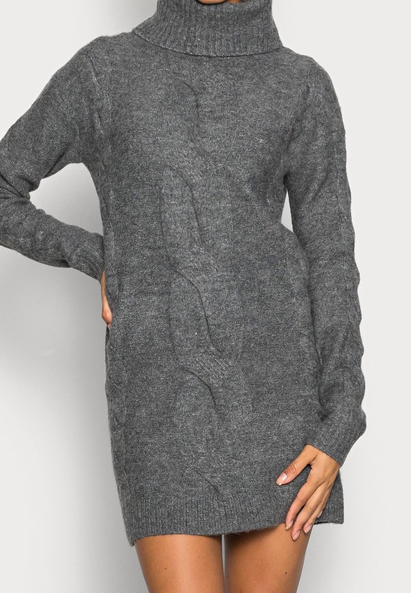 Femme portant une robe pull col roulé en maille torsadée grise ajustée, avec des manches longues, se tenant avec une main sur la hanche et l'autre reposant sur la cuisse.