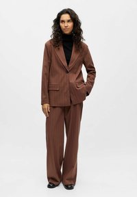 Femme aux cheveux bouclés portant un costume marron à rayures et un col roulé noir, se tenant avec une main dans sa poche contre un fond uni.