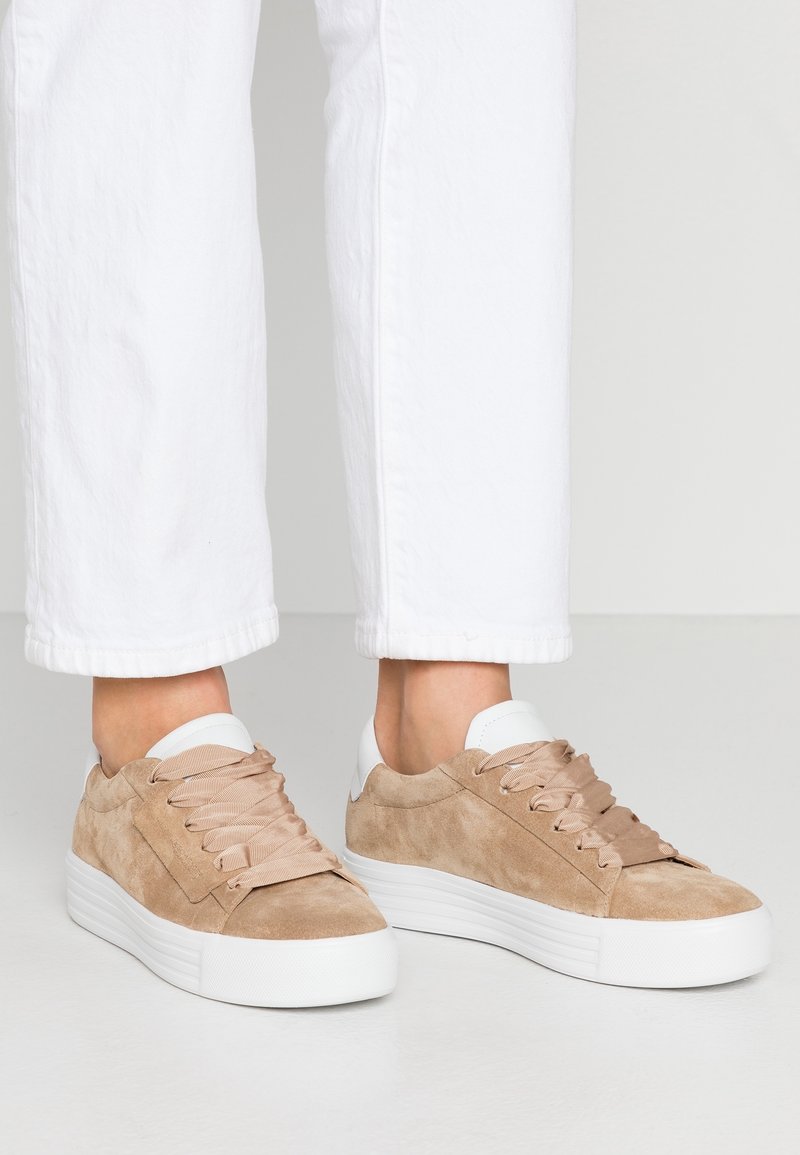 Des pieds portant des baskets en daim beige avec de larges lacets beiges et de épaisses semelles blanches, associés à un pantalon blanc sur fond uni.