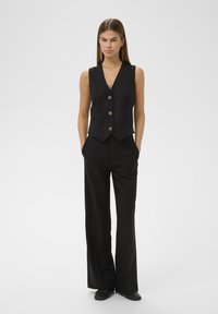 Juodas be rankovių liemenė su giliu V-kaklu ir penkiomis auksinėmis sagomis, derinama su aukštu liemeniu juodomis kelnėmis ir juodais plokščiapadžiais batais.