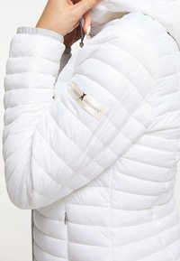 Osoba w białej pikowanej kurtce z logo Frieda & Freddies New York na rękawie, poprawiająca kaptur ręką przy twarzy.