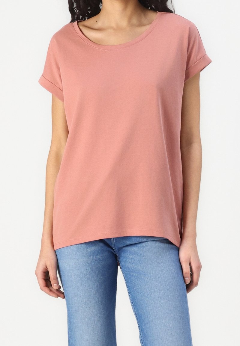 Femme portant un t-shirt rose ample à manches courtes et un jean bleu clair sur un fond blanc uni.