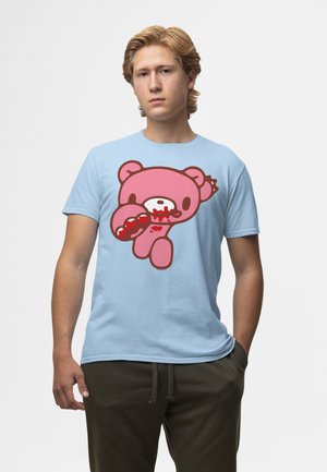 Ung mand iført en lyseblå t-shirt med en grafik af en lyserød bamse med blod på munden og poten, stående mod en ensfarvet baggrund.