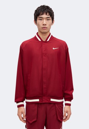 Jeune homme portant une veste de survêtement Nike rouge et un pantalon assorti avec des bandes blanches aux poignets et à la taille, debout devant un fond uni.