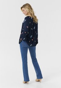 Blouse marine avec des motifs brodés colorés, coupe décontractée, manches longues ; associée à un jean flare bleu clair et des talons beiges.