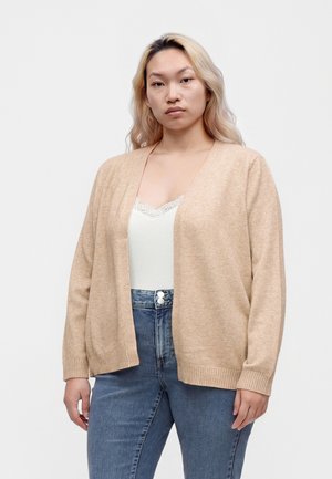 Ung kvinde med langt blondt hår iført beige åben cardigan, hvid top med blonderkant og blå høj-taljede jeans mod en ensfarvet baggrund.