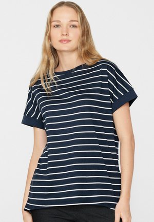 Junge Frau mit langen blonden Haaren, die ein dunkelblaues Kurzarmshirt mit weißen horizontalen Streifen trägt, steht vor weißem Hintergrund.