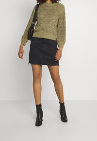 Suéter de punto verde oliva con patrón texturizado, combinado con una falda corta de mezclilla negra y botas de tobillo negras. Se incluye un bolso negro de hombro.