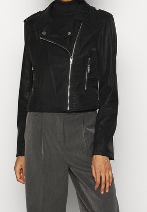 Woman wearing a black zipped biker jacket over a black turtleneck and gray pleated trousers, standing against a plain background.