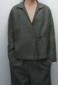 Veste gris foncé en mélange de lin avec une coupe décontractée, devant ouvert, col pointu et détail de lien à la taille. Surface texturée et poches latérales.