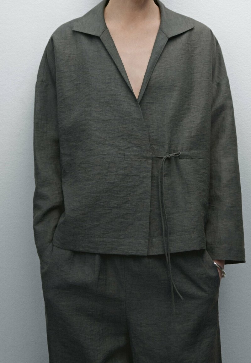 Veste gris foncé en mélange de lin avec une coupe décontractée, devant ouvert, col pointu et détail de lien à la taille. Surface texturée et poches latérales.