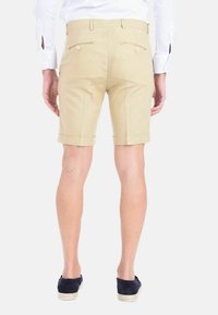 Pantaloni corti beige in lino con risvolti, dotati di due tasche posteriori con chiusura a bottone, indossati con scarpe scure, visti da dietro.