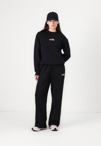 Svart hettegenser og matchende bukser, laget av myk stoff, med en liten flerfarget logo på brystet. Båret med en svartCaps og sneakers.