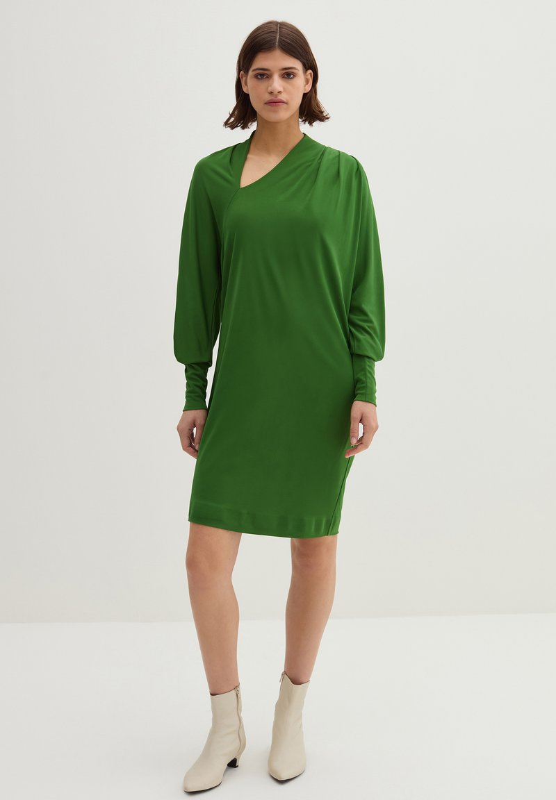 Groene jurk met lange, opgerichte mouwen, V-halsontwerp, gemaakt van een gladde stof. Draag met beige enkellaarsjes, tegen een neutrale achtergrond.