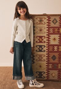 Cardigan à manches longues côtelé crème avec boutons, associé à un jean en denim foncé à jambes larges. La fille porte des chaussures blanches avec un motif noir.