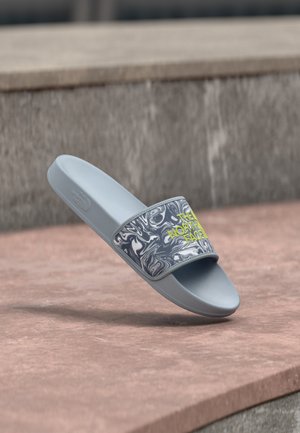 Sandalo da scivolo azzurro chiaro con cinturino motivo marmo e logo giallo "The North Face" che poggia su una superficie in pietra.