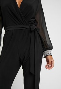 Svart jumpsuit med djup V-ringning och omslagdesign. Genomskinliga långärmar med manschett dekorerad med rhinestones. Knuten midjedetalj.