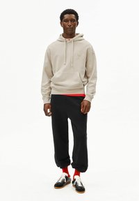 Beige hoodie met trekkoorden en voorzak, gecombineerd met zwarte joggingbroek. Model draagt rode sokken en zwarte sneakers met witte accenten.