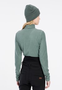 Haut à manches longues vert côtelé avec un col montant, associé à un pantalon noir taille haute. Un bonnet en tricot vert assorti complète la tenue.